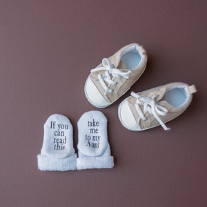 May include: A pair of white baby socks with the text "If you can read this" and "take me to my Aunt" on them.  Next to the socks are a pair of beige baby shoes with white laces.