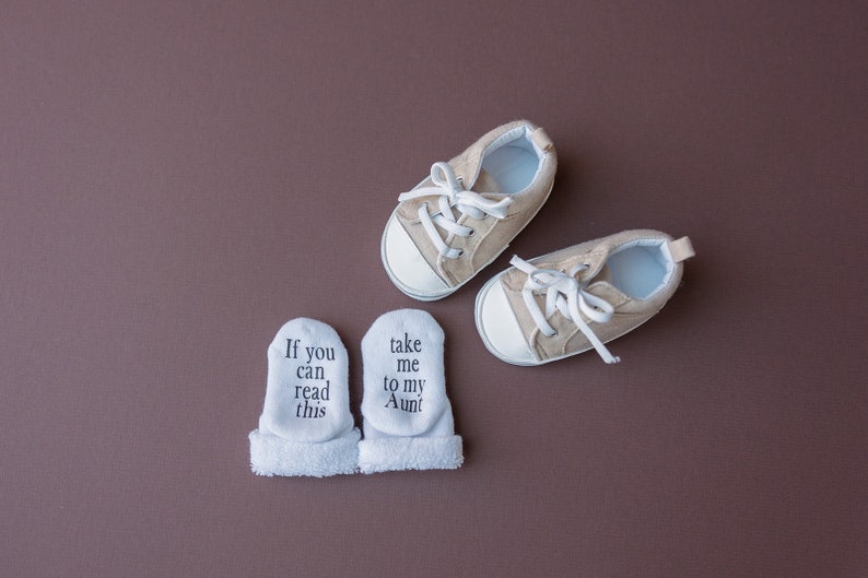 May include: A pair of white baby socks with the text "If you can read this" and "take me to my Aunt" on them.  Next to the socks are a pair of beige baby shoes with white laces.