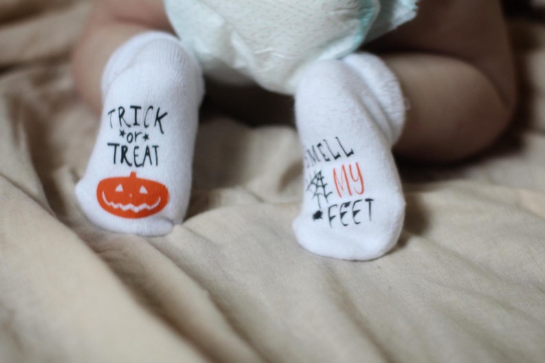 May include: A pair of white baby socks with fun Halloween-themed designs. One sock says "Trick or Treat" with a pumpkin face, and the other says "Smell My Feet" with a spider web.