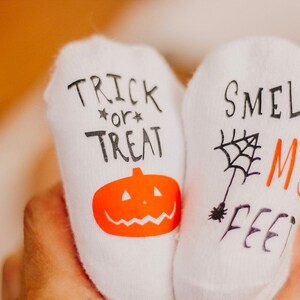 May include: A pair of white socks with Halloween-themed designs. One sock says "Trick or Treat" with a pumpkin and the other says "Smell My Feet" with a spider web.