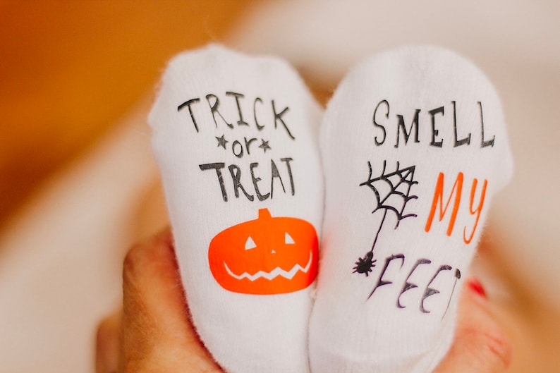 May include: A pair of white socks with Halloween-themed designs. One sock says "Trick or Treat" with a pumpkin and the other says "Smell My Feet" with a spider web.