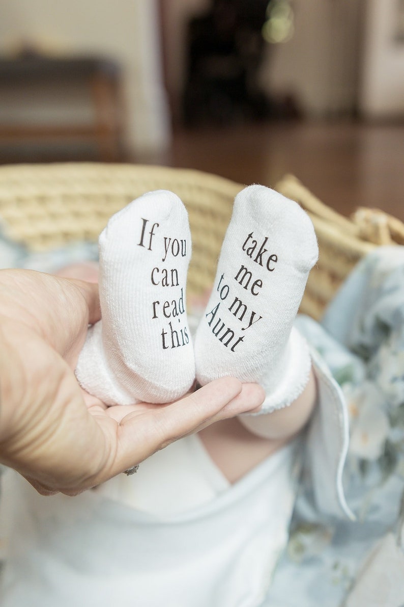 May include: A pair of white baby socks with black text. One sock reads "If you can read this" and the other sock reads "Take me to my Aunt".