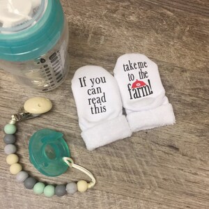 K&ouml;nnte beinhalten: Ein Paar wei&szlig;e Baby-Sch&uuml;hchen mit schwarzem Text. Ein Sch&uuml;hchen sagt "Wenn du das lesen kannst" und das andere sagt "Bring mich zum Bauernhof!" mit einem roten Scheunendach-Symbol.