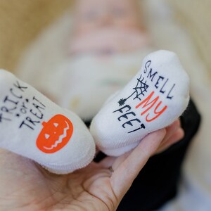 May include: A pair of white baby socks with Halloween-themed designs. One sock says "Trick or Treat" with an orange pumpkin and the other says "Smell My Feet" with a spider web.