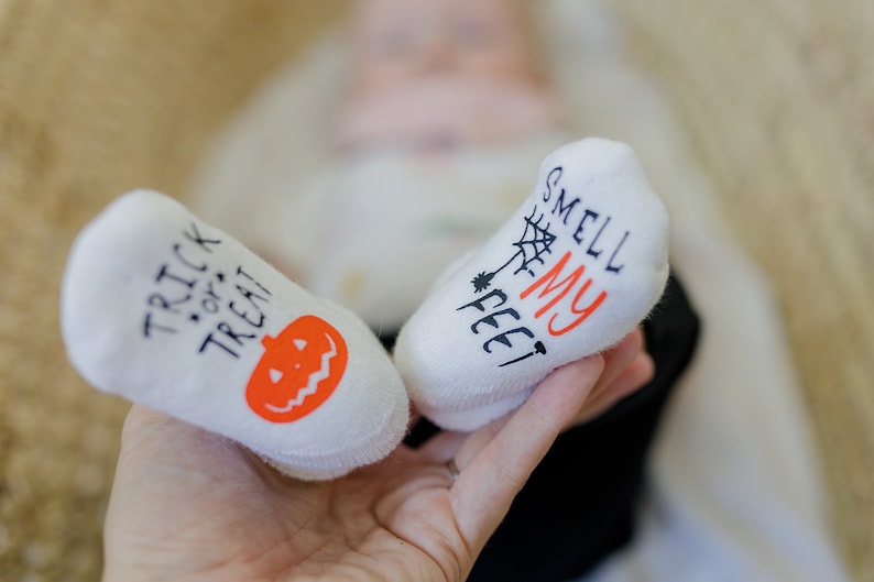 May include: A pair of white baby socks with Halloween-themed designs. One sock says "Trick or Treat" with an orange pumpkin and the other says "Smell My Feet" with a spider web.