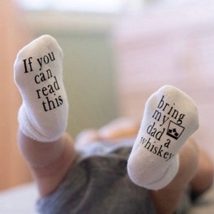 May include: A pair of white baby socks with black text. One sock reads "If you can read this" and the other sock reads "bring my dad a whiskey".