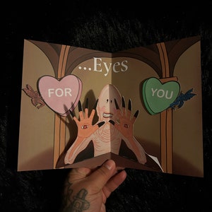 May include: A pop-up greeting card featuring a pale figure with long black nails and eyes in its palms. The card has the text "...Eyes" and heart-shaped candies that read "FOR" and "YOU".