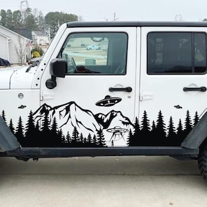 May include: A white Jeep Wrangler with a black vinyl decal on the side. The decal features a mountain range, pine trees, and flying saucers.