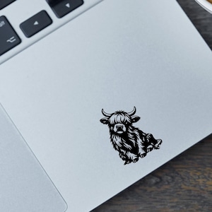 May include: A black vinyl decal featuring a shaggy Highland cow illustration applied to a silver laptop surface. The text above reads: VALDON IMAGES, Unique Customizable Vinyl Decals!, different colors & sizes to choose from.