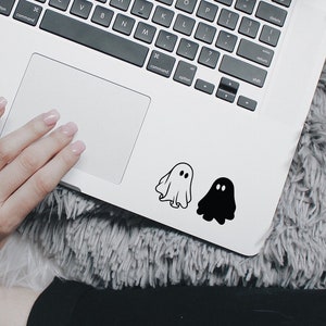 Puede incluir: Dos pegatinas de fantasmas en un portátil. Un fantasma es blanco y el otro es negro.