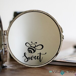 Op de afbeelding: Een zilveren compact spiegel met een zwart-witte bij-illustratie en de tekst "Sweet" aan de binnenkant van de spiegel.