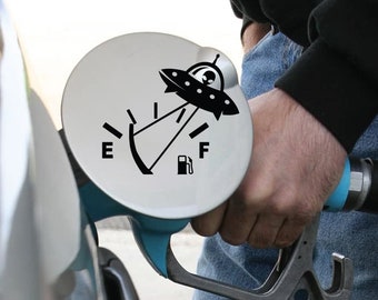 May include: A gas tank cap with a black and white UFO decal. The gas gauge is pointing to 'F' for full. The decal reads 'E' for empty and 'F' for full.
