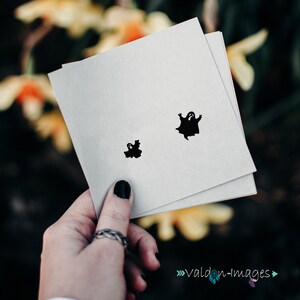 May include: A white card with two black ghost silhouettes. The larger ghost is in the centre of the card, and the smaller ghost is in the bottom left corner.