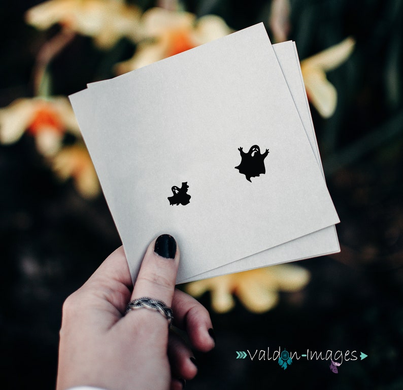 May include: A white card with two black ghost silhouettes. The larger ghost is in the centre of the card, and the smaller ghost is in the bottom left corner.