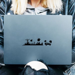 May include: A black laptop computer with a Halloween-themed sticker on the lid. The sticker depicts a graveyard scene with a spooky tree, tombstones, ghosts, and a car.