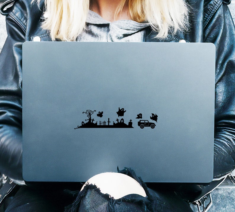 May include: A black laptop computer with a Halloween-themed sticker on the lid. The sticker depicts a graveyard scene with a spooky tree, tombstones, ghosts, and a car.