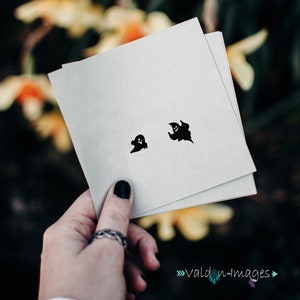 May include: Two black ghost stickers on a white card. The card is being held by a hand with black nail polish.