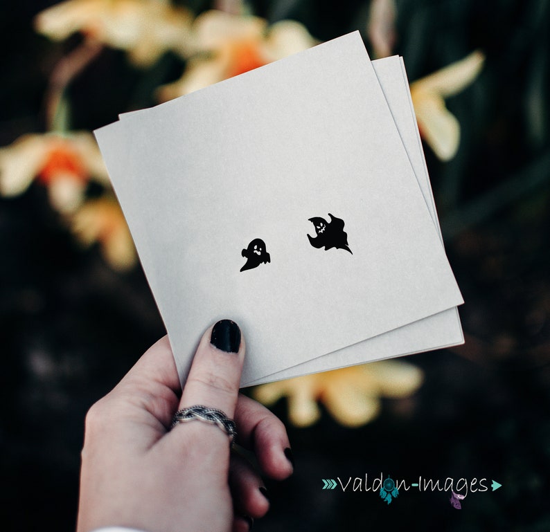 May include: Two black ghost stickers on a white card. The card is being held by a hand with black nail polish.