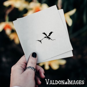 May include: A black and white silhouette of a dragon flying over a mountain range. The image is printed on a white card.