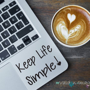 May include: A laptop computer with a silver finish and a black keyboard. The laptop has a sticker on the touchpad that says "Keep Life Simple!" with a small heart. A cup of coffee with latte art is on a wooden table next to the laptop.
