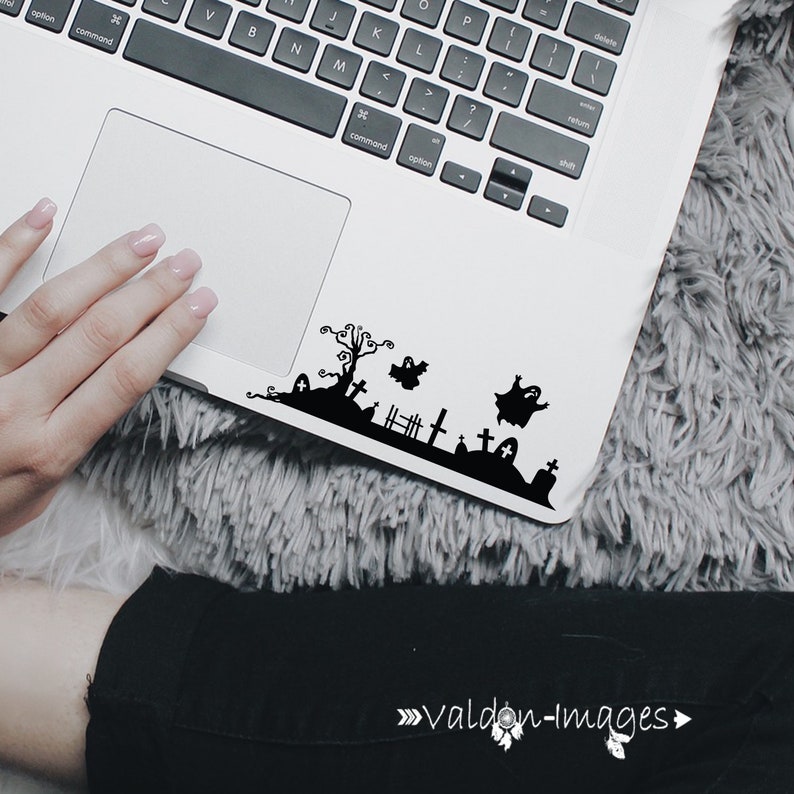 May include: A white laptop with a black Halloween-themed sticker on the bottom right corner. The sticker features a spooky graveyard scene with a tree, ghosts, and tombstones.
