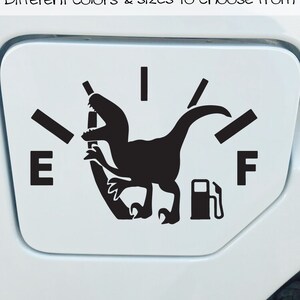 May include: A black and white vinyl decal of a dinosaur with a gas pump in its mouth. The dinosaur is positioned on a gas gauge with the letters "E" and "F" on either side. The decal is designed to be placed on a car's gas tank.