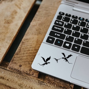 May include: A silver laptop computer with a black keyboard and a black trackpad. There are three black dragon stickers on the trackpad.
