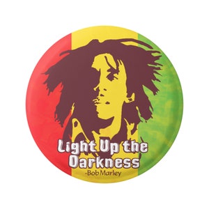Puede incluir: Un botón redondo con fondo rojo, amarillo y verde. El botón presenta una silueta de Bob Marley con el texto "Light Up the Darkness -Bob Marley".