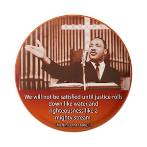 Puede incluir: Un botón redondo con una foto de Martin Luther King Jr. hablando desde un podio. La cita "We will not be satisfied until justice rolls down like water and righteousness like a mighty stream. -Martin Luther King, Jr." está impresa debajo de la foto.