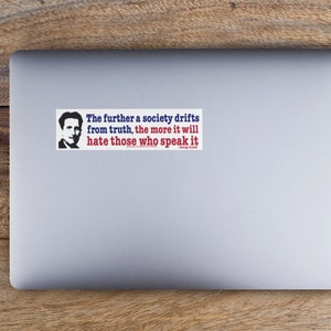 May include: A rectangular sticker on a laptop with a quote by George Orwell. The text reads: "The further a society drifts from truth, the more it will hate those who speak it." The sticker has a portrait of a man.