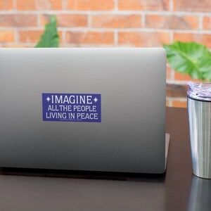 May include: A blue sticker with white text on a silver laptop. The sticker says "*IMAGINE* ALL THE PEOPLE LIVING IN PEACE".