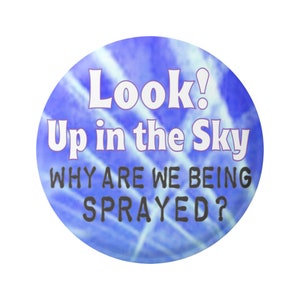Puede incluir: Un botón azul y blanco tie-dye con el texto "Look! Up in the Sky WHY ARE WE BEING SPRAYED?"