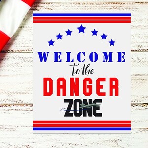 May include: A white sign with red, white, and blue stripes and stars. The sign says "Welcome to the Danger Zone" with a black and gray airplane graphic.