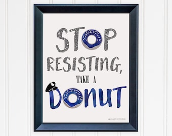 May include: A framed print with a white background and black text that reads "Stop Resisting, Take a Donut". The text is in a glittery silver font. A blue donut with white sprinkles is depicted in the text. A blue police officer's hat is on top of the "D" in "Donut".