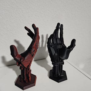 May include: Two 3D printed robotic hands, one red and one black, are posed with their palms facing up. The hands are on a white surface and are on hexagonal bases.
