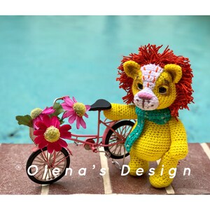 May include: A handmade yellow crochet lion with a red mane and a green scarf stands next to a miniature pink bicycle filled with pink and yellow flowers. The lion has a white and orange face. The text "Olena's Design" is visible.
