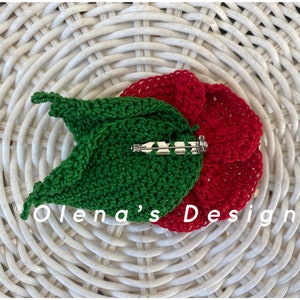 May include: A handmade crochet flower pin with a red flower and green leaf design. The pin features a silver-colored clasp on the back. The text "Olena's Design" is visible on the bottom left.