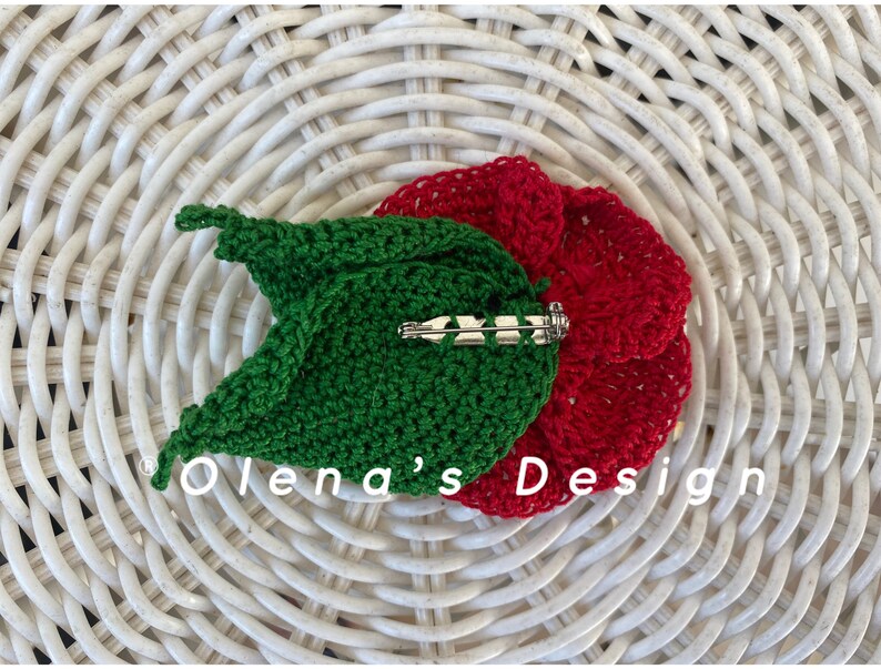 May include: A handmade crochet flower pin with a red flower and green leaf design. The pin features a silver-colored clasp on the back. The text "Olena's Design" is visible on the bottom left.