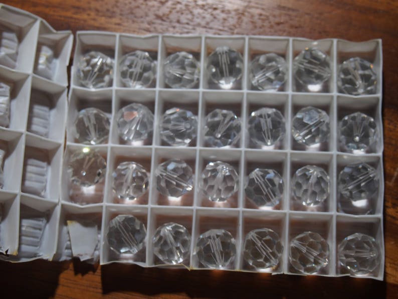 May include: A close-up shot of clear, faceted crystal beads arranged in a white compartmentalized box. The beads are spherical and reflect light, creating a sparkling effect. The box is open, revealing multiple beads.