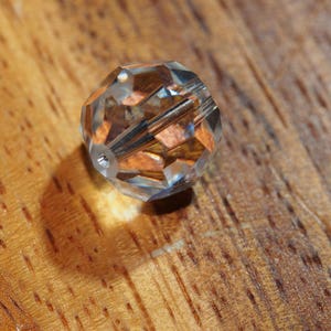 May include: A clear, faceted crystal bead with a hole running through the center. The bead reflects light, creating a sparkling effect. The bead is resting on a wooden surface.