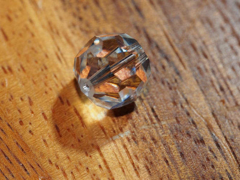 May include: A clear, faceted crystal bead with a hole running through the center. The bead reflects light, creating a sparkling effect. The bead is resting on a wooden surface.