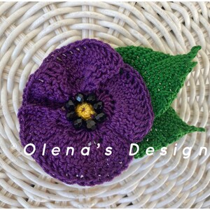 May include: A handmade crochet flower with a purple bloom, a yellow center, and black bead accents. Two green crochet leaves are attached to the flower. The text "Olena's Design" is visible on the image.