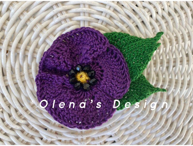 May include: A handmade crochet flower with a purple bloom, a yellow center, and black bead accents. Two green crochet leaves are attached to the flower. The text "Olena's Design" is visible on the image.