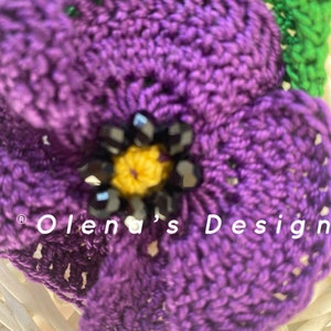May include: Close-up of a handmade crochet flower in shades of purple, with a yellow center and black bead accents. The flower is part of a larger design, with the text "Olena's Design" visible.
