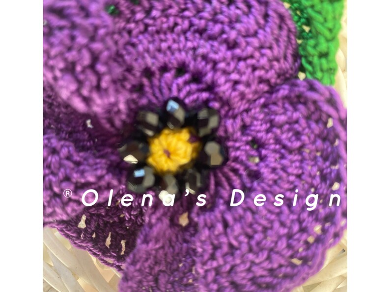 May include: Close-up of a handmade crochet flower in shades of purple, with a yellow center and black bead accents. The flower is part of a larger design, with the text "Olena's Design" visible.
