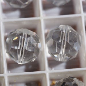May include: Close-up of clear, faceted crystal beads. The beads are round with multiple flat sides, creating a sparkling effect. They are arranged in a white, compartmentalized tray, suggesting organization and storage.