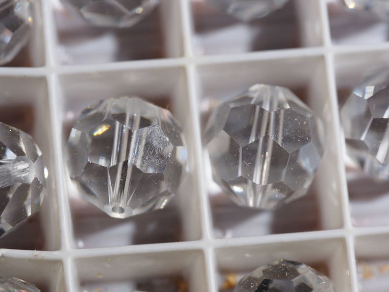 May include: Close-up of clear, faceted crystal beads. The beads are round with multiple flat sides, creating a sparkling effect. They are arranged in a white, compartmentalized tray, suggesting organization and storage.
