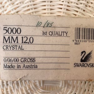 May include: A rectangular, off-white label with black text and a barcode. The label reads "5000 MM 12,0 CRYSTAL" and "Made in Austria." The Swarovski logo is visible. The label is on a white woven surface.