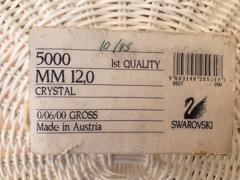 May include: A rectangular, off-white label with black text and a barcode. The label reads "5000 MM 12,0 CRYSTAL" and "Made in Austria." The Swarovski logo is visible. The label is on a white woven surface.