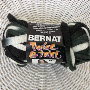 May include: A skein of Bernat Twist & Twirl yarn in shades of black, green, and white. The label indicates the yarn is 100% acrylic and weighs 3.5 oz. The yarn is displayed on a white woven surface.